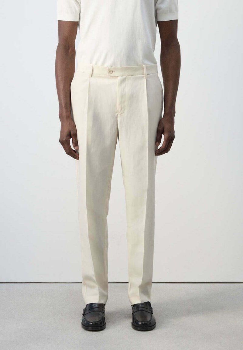 Homme portant un pantalon plissé de couleur crème, une chemise blanche à manches courtes et des mocassins noirs, debout devant un fond uni.