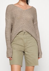 Hellbrauner Strickpullover mit V-Ausschnitt, langen Ärmeln und lockerer Passform, kombiniert mit olivgrünen Jeansshorts mit fransigem Saum.