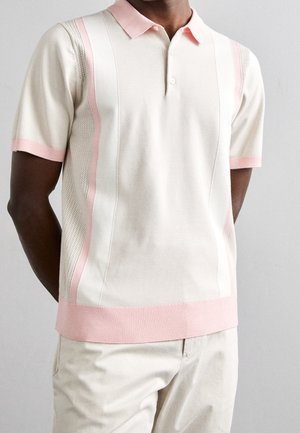 Homme portant un polo à manches courtes de couleur crème avec des bordures rose clair, associé à un pantalon crème, debout devant un fond gris uni.