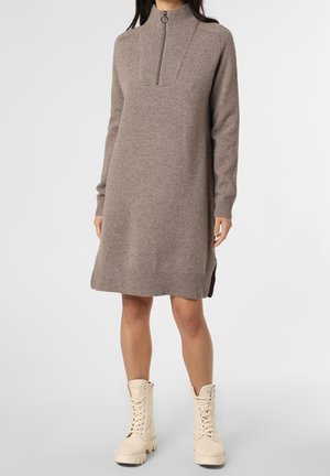 Femme portant une robe en maille taupe à manches longues avec une fermeture éclair sur le devant et des bottes crème à lacets, debout sur un fond uni.