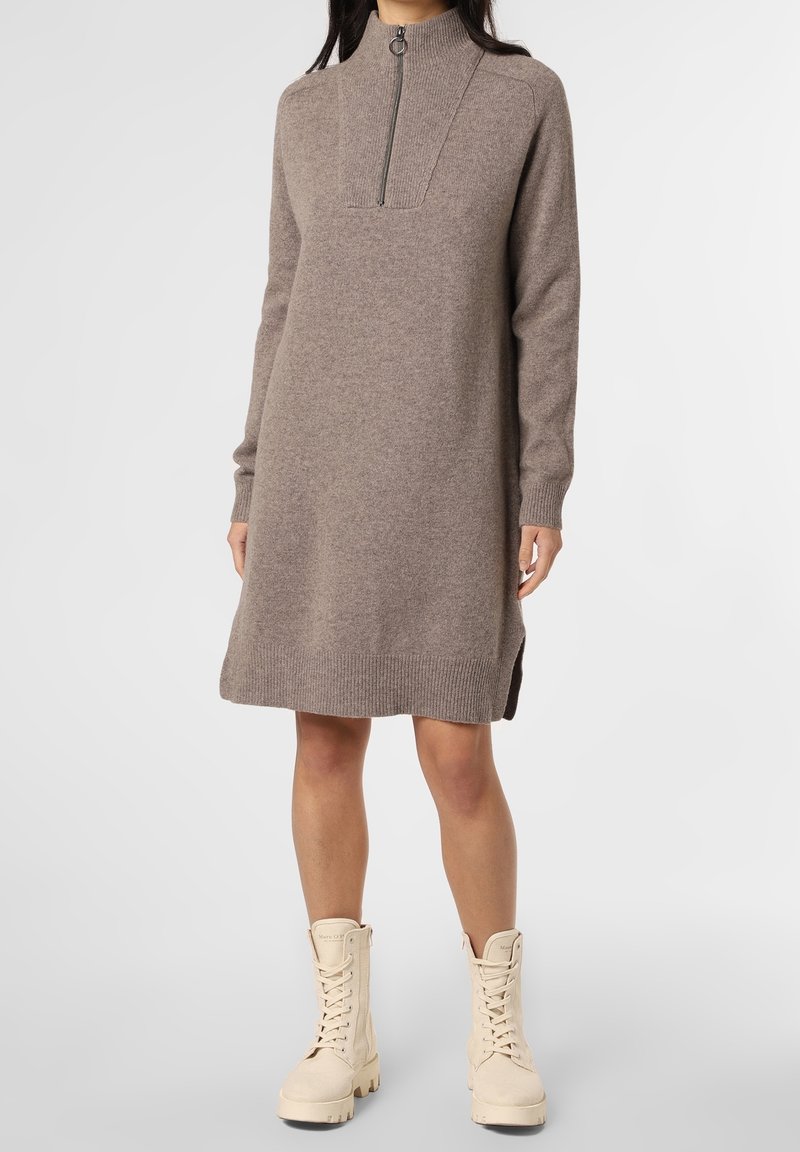 Femme portant une robe en maille taupe à manches longues avec une fermeture éclair sur le devant et des bottes crème à lacets, debout sur un fond uni.