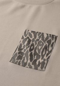 Beige shirt with a textured black and gray leopard print pocket, featuring a rectangular shape and subtle stitching detail.