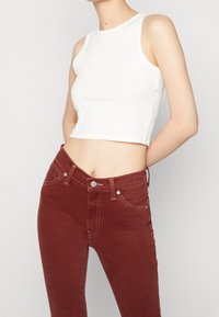 Mujer vistiendo un top corto sin mangas blanco y jeans de tiro alto de color óxido, de pie con las manos detrás de la espalda sobre un fondo liso.