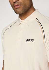 Világos bézs polo ing klasszikus gallérral, fekete "BOSS" logóval, perforált részletekkel a galléron és vékony kiemelő csíkokkal.