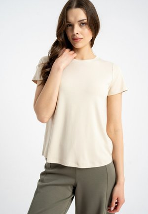 Kobieta z długimi brązowymi włosami, ubrana w beżową koszulę z krótkim rękawem i oliwkowe zielone spodnie, stojąca na jednolitym jasnym tle.