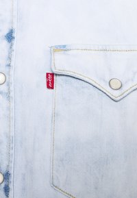 Camicia in denim azzurro chiaro con tasca frontale, cuciture gialle, chiusura a bottoni e etichetta rossa Levi's sulla tasca.