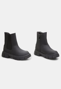 Botas de tobillo negras con suela gruesa, material de textura suave, paneles laterales elásticos y cierre de cremallera en un lado.