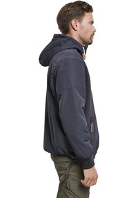 Chaqueta ligera azul marino con capucha, cremallera frontal y cordón en el dobladillo. Cuenta con puños elásticos y un bolsillo lateral con cremallera.