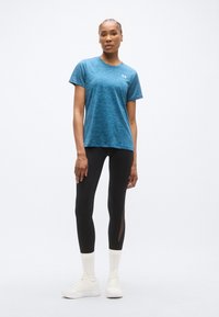 Μπλε αθλητική μπλούζα κοντομάνικη με υφή, σε συνδυασμό με μαύρο κολάν που έχει πλαϊνή λεπτομέρεια από δίχτυ, και λευκά αθλητικά παπούτσια.