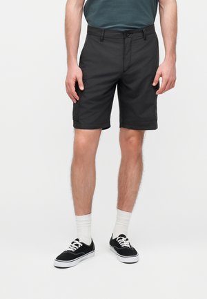 Άνδρας που φοράει μαύρα cargo σορτς, λευκές κάλτσες και μαύρα αθλητικά, στέκεται μπροστά σε ένα απλό φωτεινό φόντο.