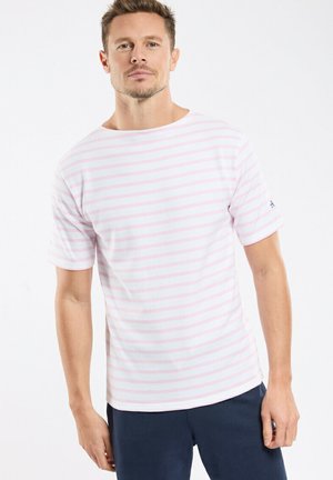 Homme portant une chemise à manches courtes rayée horizontalement de blanc et de rose clair, et un pantalon bleu foncé, debout devant un fond blanc uni.