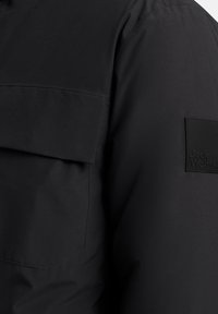 Veste noire au toucher lisse, dotée d'une poche sur la poitrine et d'une étiquette logo en caoutchouc. Le design est moderne et fonctionnel.