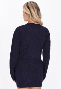 Neulepuseromekko, tummansininen metallilangoilla. Ominaisuuksiin kuuluu pitkät hihat, teksturoitu palmikkoneulekuvio, hyvin istuva vyötärö ja pyöreä kaula-aukko.