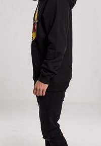 Homme debout de profil portant un sweat à capuche noir avec un motif coloré et un jean noir déchiré, sur un fond clair uni.