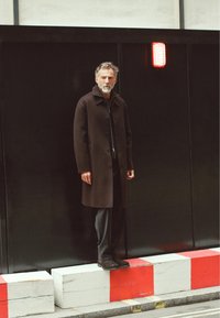 Long brown coat with a collar and buttons, worn over a black top and gray trousers. The figure stands on a white block with a red stripe.