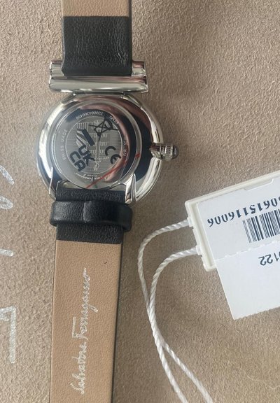 Black leather strap with silver and tan accents, round silver case, black dial with white markings, and a tag attached.