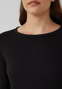 Zwarte ribbel langek mouwtop met een ronde halslijn en een klein, reliëflogo op het midden van de borst. Soepele textuur.