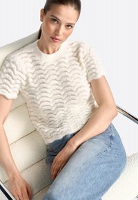Kortærmet cremefarvet sweater med bølget tekstur, lavet af blødt materiale. Sammen med lyseblå denimjeans, siddende på en hvid stol.