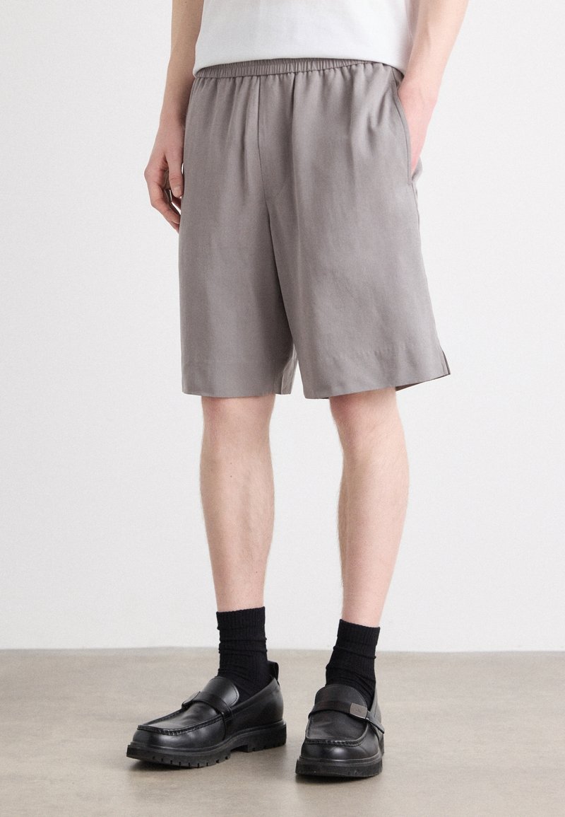 Personne portant un short gris ample avec des poches latérales, des chaussettes noires et des mocassins en cuir noir, debout sur un sol lisse contre un mur blanc.