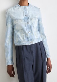 Ljust blå denimjacka med en kortad design, prydd med broderade mönster, silverknappar och en rund halsringning.