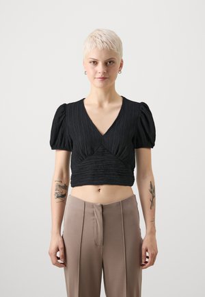 Black cropped top with a textured finish, deep V-neck, and puff sleeves. Paired with light beige high-waisted trousers featuring pleats.