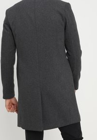 Grauer Wollmantel mit tailliertem Schnitt, Revers mit Kerbkragen und einem einzelnen Schlitz hinten, mit langen Ärmeln und sauberem Abschluss.