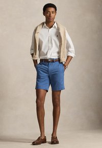 Młody mężczyzna ubrany w białą koszulę na guziki, beżowy sweter zarzucony na ramiona, niebieskie spodenki, brązowy pasek i brązowe mokasyny.