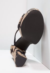Sandalia negra de tacón alto con suela texturizada. La parte superior presenta motivos florales metálicos en dorado y beige, con una fina correa en el tobillo.