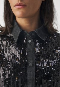 Camicia in denim nero decorata con paillettes di varie sfumature, dotata di una chiusura con bottoni e colletto a punta, che presenta una superficie testurizzata.