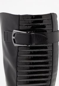 Botte en cuir noir avec un panneau en cuir laqué à rayures verticales, une accent en boucle en argent et une texture lisse.