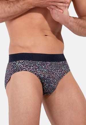 Männlicher Oberkörper mit dunklen Slips mit floralem Muster und breitem schwarzem Bund, die Hände sind vor der Brust zusammengehalten.