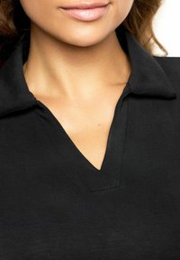 Parte inferiore del viso e collo di donna che indossa una camicia nera con colletto e scollatura a V su uno sfondo bianco.