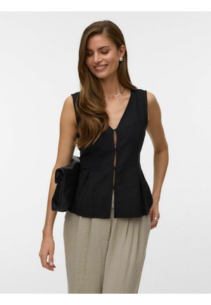 Kvinde med langt brunt hår iført sort ærmeløs knapbluse, beige bukser, perleøreringe, sølvsmykke og holder en sort clutch taske.