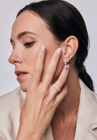 Złote kolczyki z trzema przezroczystymi kamieniami, teksturowany złoty pierścień z małymi koralikami i kremowa marynarka. Palec delikatnie opiera się na policzku.