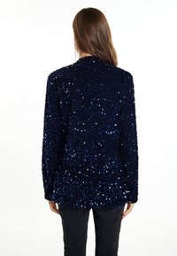 Chaqueta de lentejuelas azul marino con una superficie texturizada, que presenta un corte relajado, mangas largas y acentos reflectantes de luz dispersos.