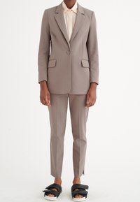 Completo marrone chiaro su misura con rever a punta, due tasche frontali e pantaloni abbinati. Indossato sopra una blusa chiara e abbinato a sandali neri.