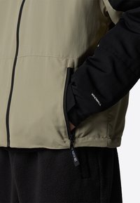 Veste beige et noire avec poche zippée et « WindWall » sur la manche, main insérée dans la poche, associée à un pantalon en polaire noir.