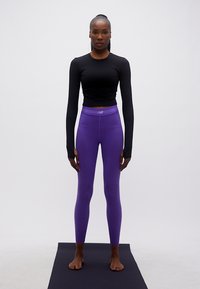 Γυναίκα όρθια, ξυπόλυτη πάνω σε μαύρο στρώμα, φοράει μαύρο τοπ με μακριά μανίκια και μοβ κολάν, κοιτάζοντας προς τα εμπρός μπροστά σε απλό φόντο.