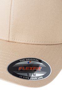 Cappello Flexfit beige realizzato in tessuto testurizzato. Presenta una corona arrotondata, visiera curvata e un adesivo logo nero e argento. Taglia S/M.