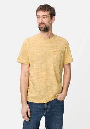 Mand med kort brunt hår og skæg iført en gul T-shirt med korte ærmer og blå jeans, stående med den ene hånd i lommen.
