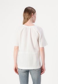 Weiße Bluse mit kurzen Ärmeln, die über strukturierte Paneele an den Schultern, ein gerafftes Rückendesign und eine leichte Taillierung verfügt.