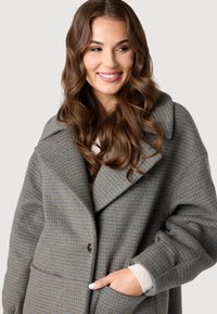 Sivý a modrý kabát s houndstooth vzorom, veľkou klopou, gombíkovým zapínaním a bočnými vreckami. Mäkka textura s uvoľneným strihom.