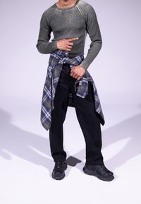 Grau gestrickter Pullover mit Rippstruktur, kombiniert mit dunklen Hosen und einem blau karierten Hemd, das um die Taille gebunden ist. Schwarze klobige Sneakers.