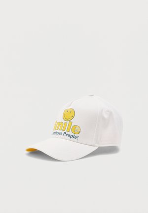 Casquette de baseball blanche avec un visage souriant jaune et le texte « Le sourire confond les gens ! » sur le devant, présentée sur un fond uni.