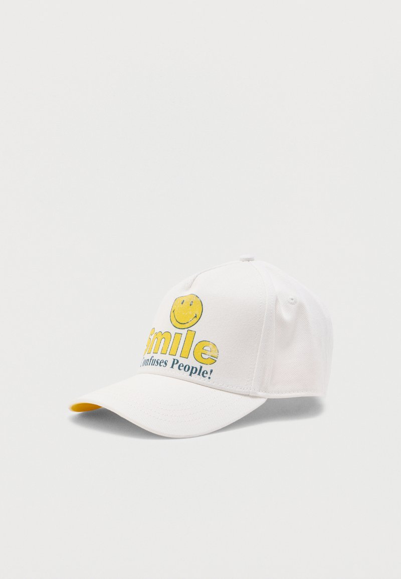 Gorra de béisbol blanca con una cara sonriente amarilla y el texto "¡La sonrisa confunde a las personas!" en la parte frontal, mostrada sobre un fondo liso.
