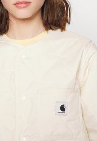 Prošívaná béžová bunda se zapínáním na patentky, obsahující náprsní kapsu a logo Carhartt. Hladká látka s jemným leskem.