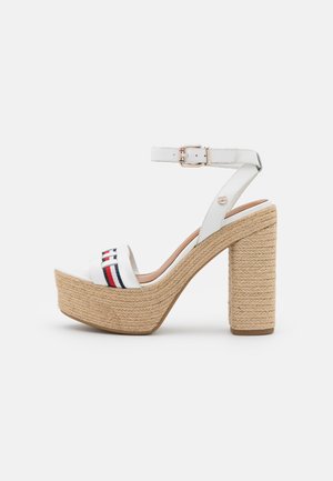 Sandale à plateforme à talon haut avec une semelle en paille tressée, une bride blanche à la cheville avec boucle, et une bride blanche à l’orteil avec des rayures rouges, blanches et bleues.