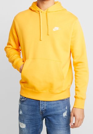 Man draagt een felgele Nike hoodie met voorzak en blauwe versleten jeans tegen een lichtgrijze achtergrond.