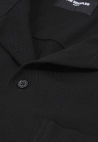 Blazer noir texturé avec un seul bouton noir, un revers cranté et une poche poitrine visible, étiquette en tissu partiellement visible à l'intérieur du col.