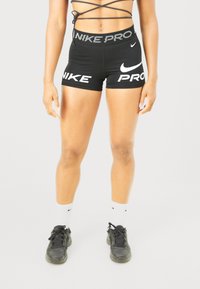 Atleta che indossa pantaloni corti neri Nike Pro, calze bianche Nike e sneakers neri, in piedi contro uno sfondo chiaro uniforme.